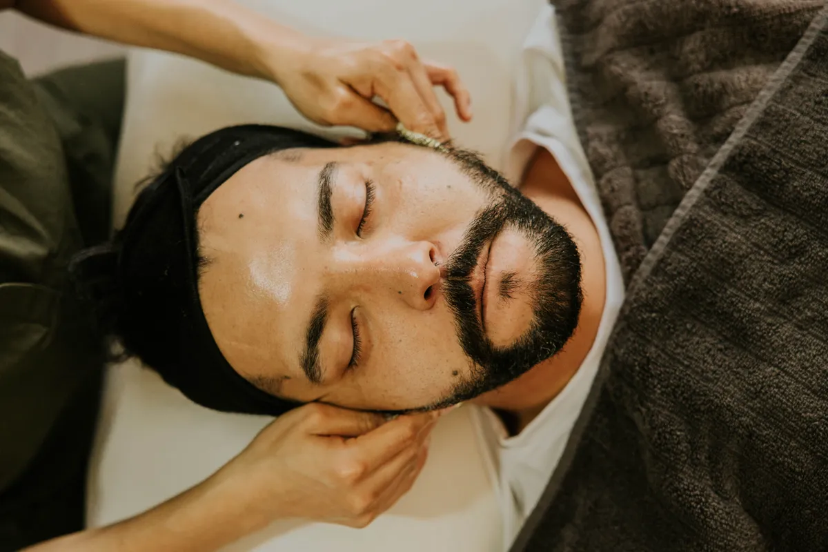 A bearded man lying on a couch with his eyes closed while a therapist's hands rest on his face