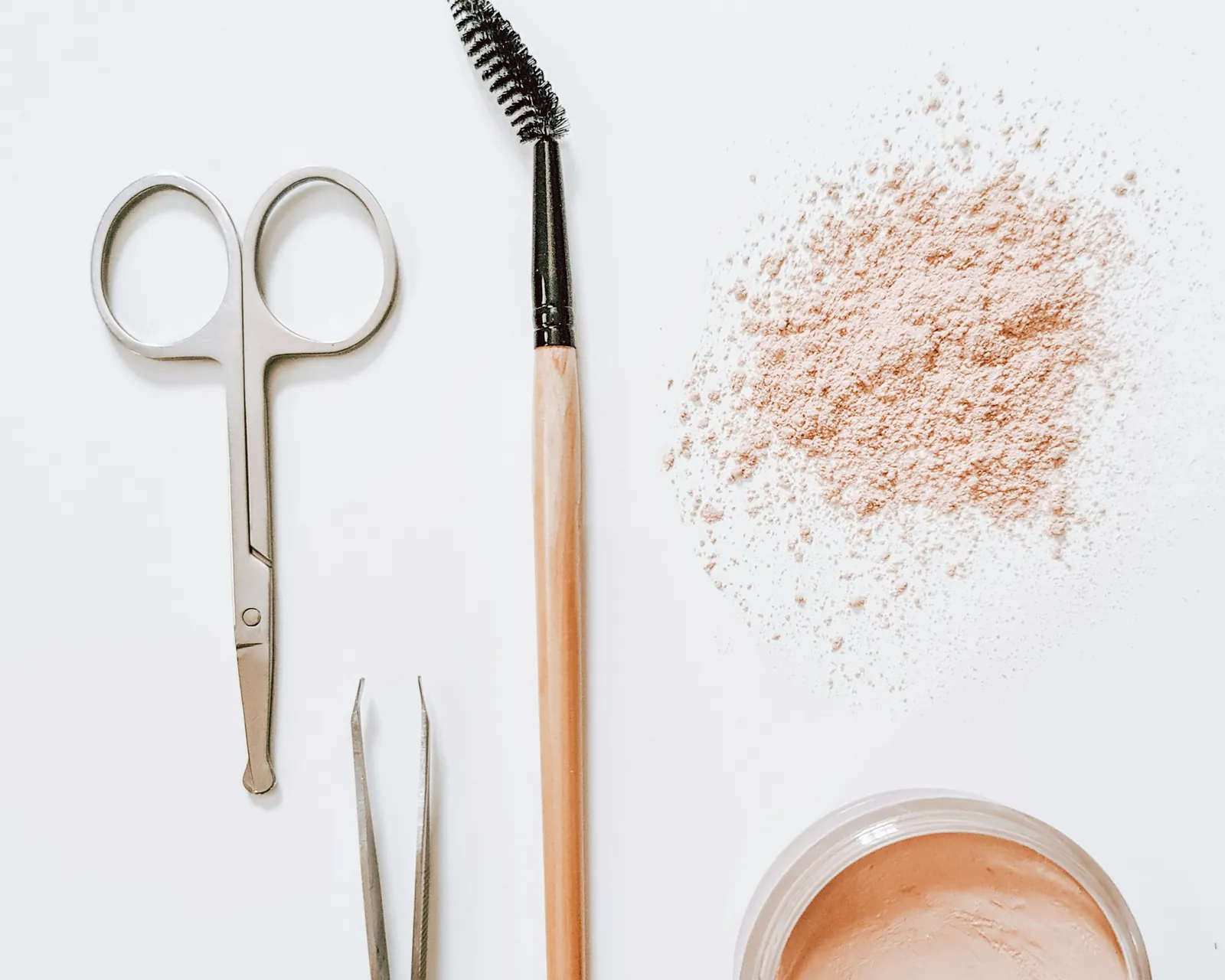 Brow scissors, a spoolie brush, tweezers and brow powder on a white surface